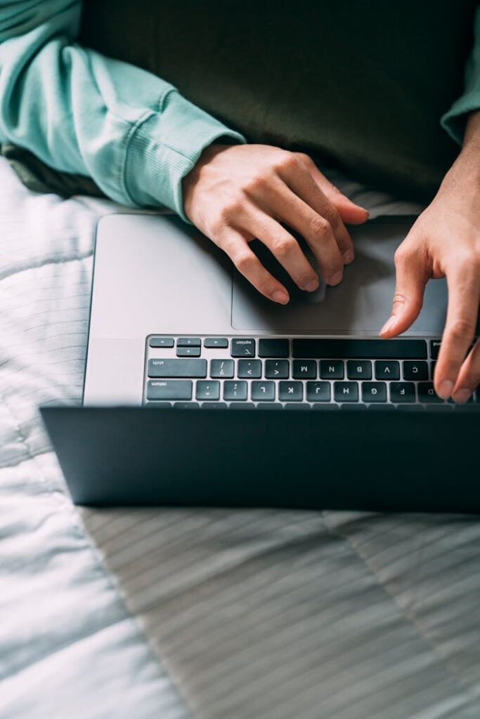 From above crop unrecognizable person using laptop while lying on comfortable bed with blanket in soft daylight